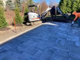 Paved driveway being installed beside shrubs, with workers and a white van in the background.