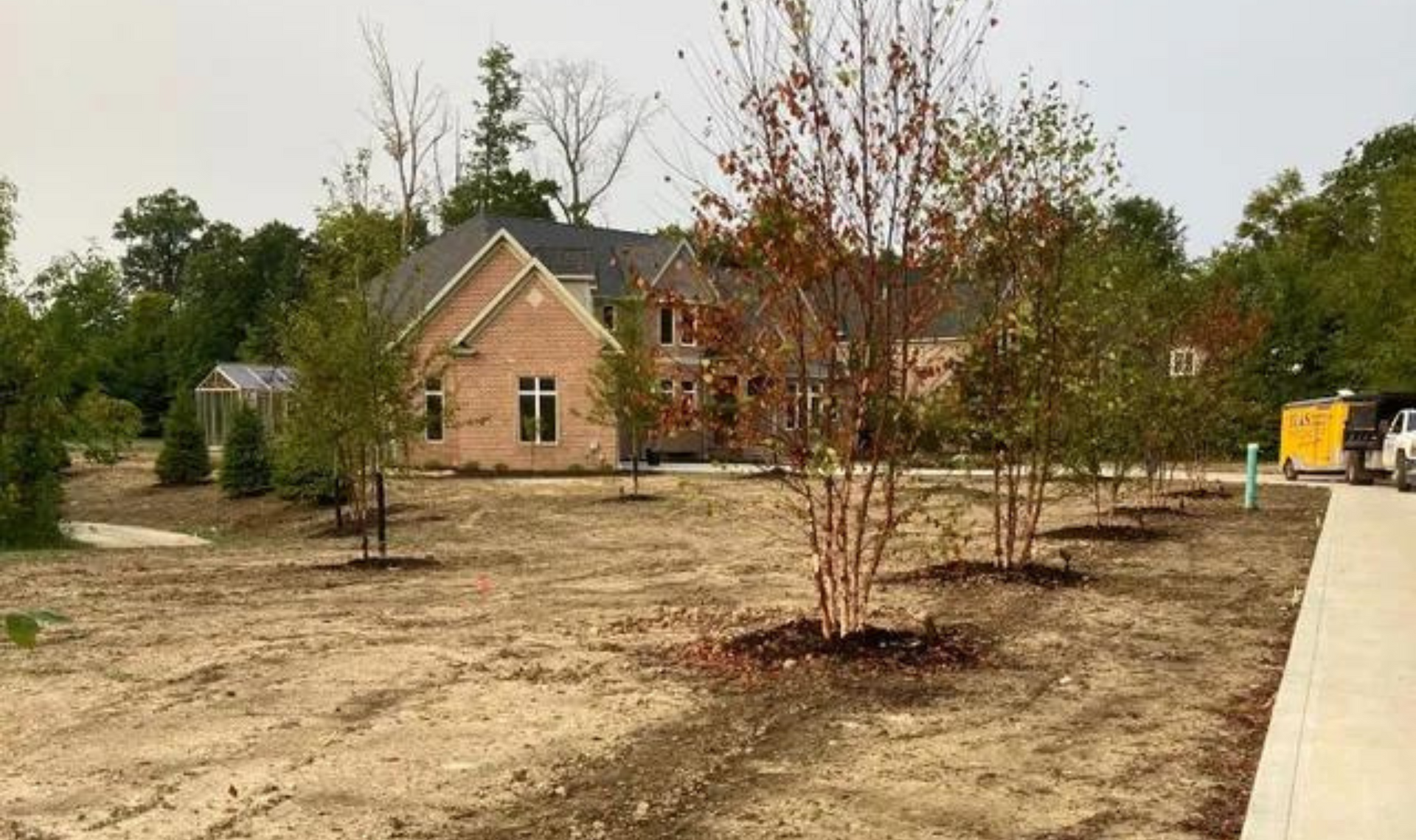 New home under construction on a bare lot with young trees and a sidewalk nearby