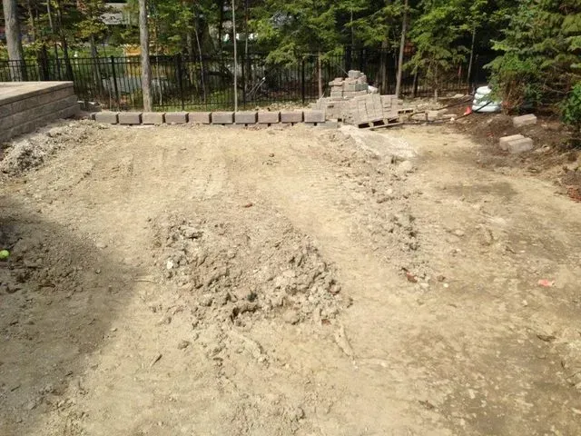 Bare dirt construction site with a sloped ground and a fence in a wooded area