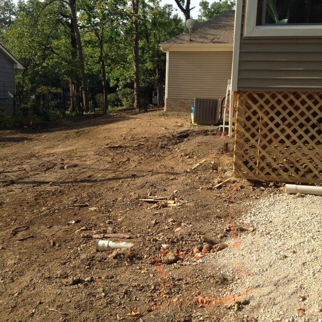 Bare dirt yard beside beige house, with gravel edge and trees in the background.
