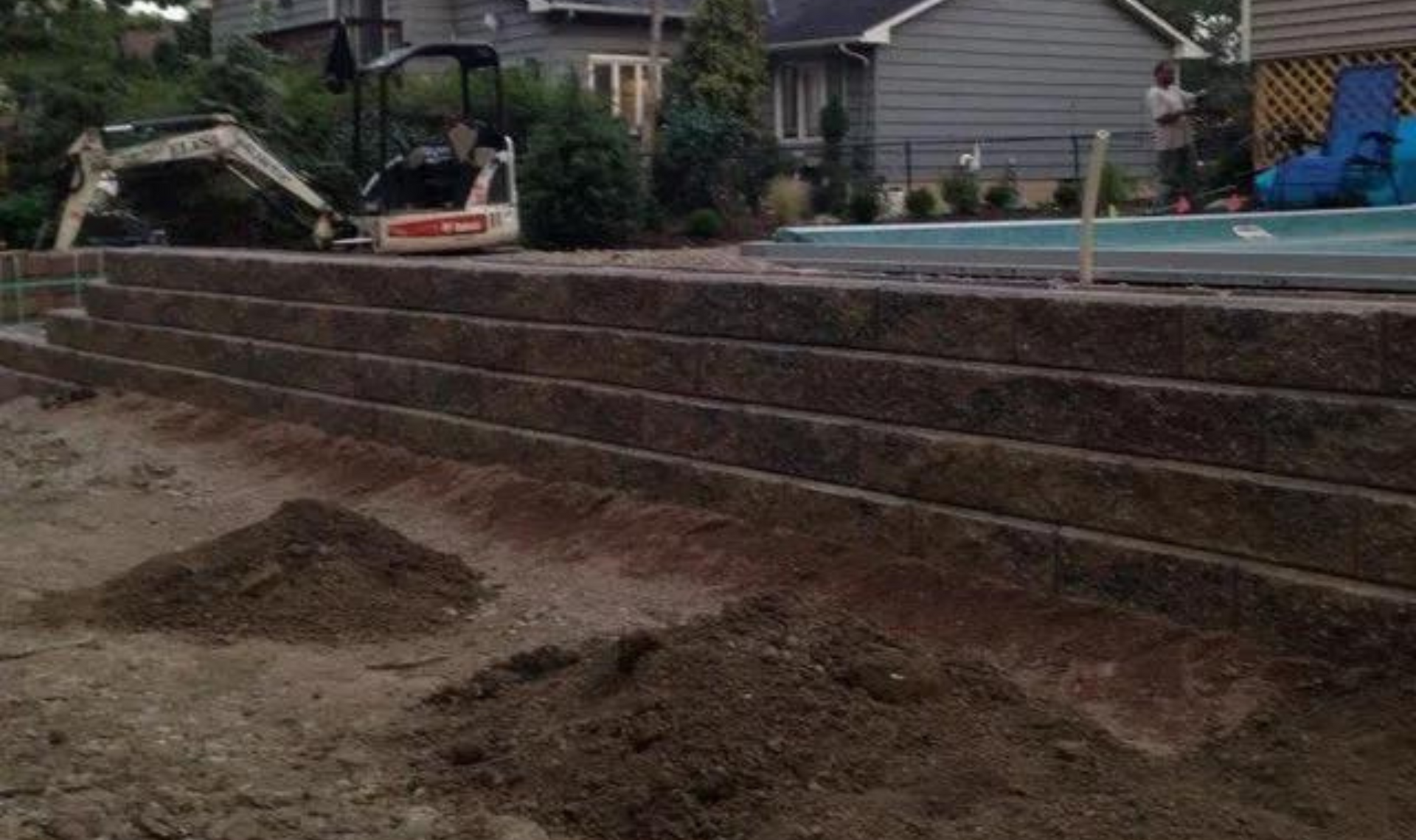 Terraced retaining wall and dirt slope beside a backyard pool, with a small excavator in the background.