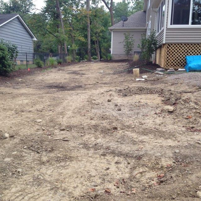 Bare dirt backyard beside two houses with a lattice deck and trees in the background