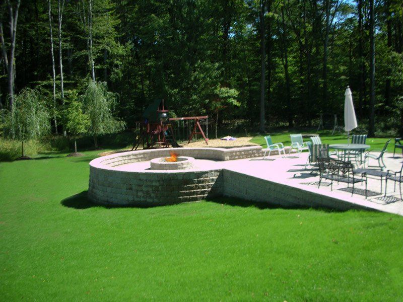 Backyard patio with stone fire pit, green lawn, outdoor seating, and trees in the background