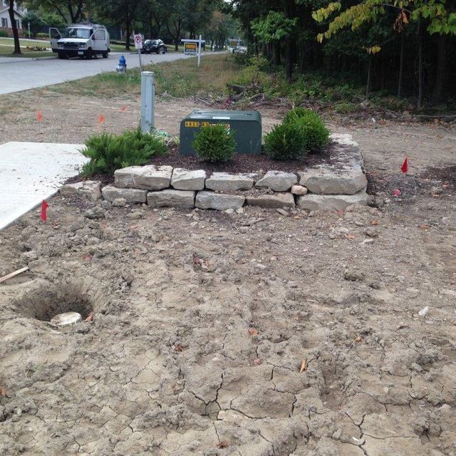 Rock garden with stacked stone border and black utility box in a dirt yard near a sidewalk