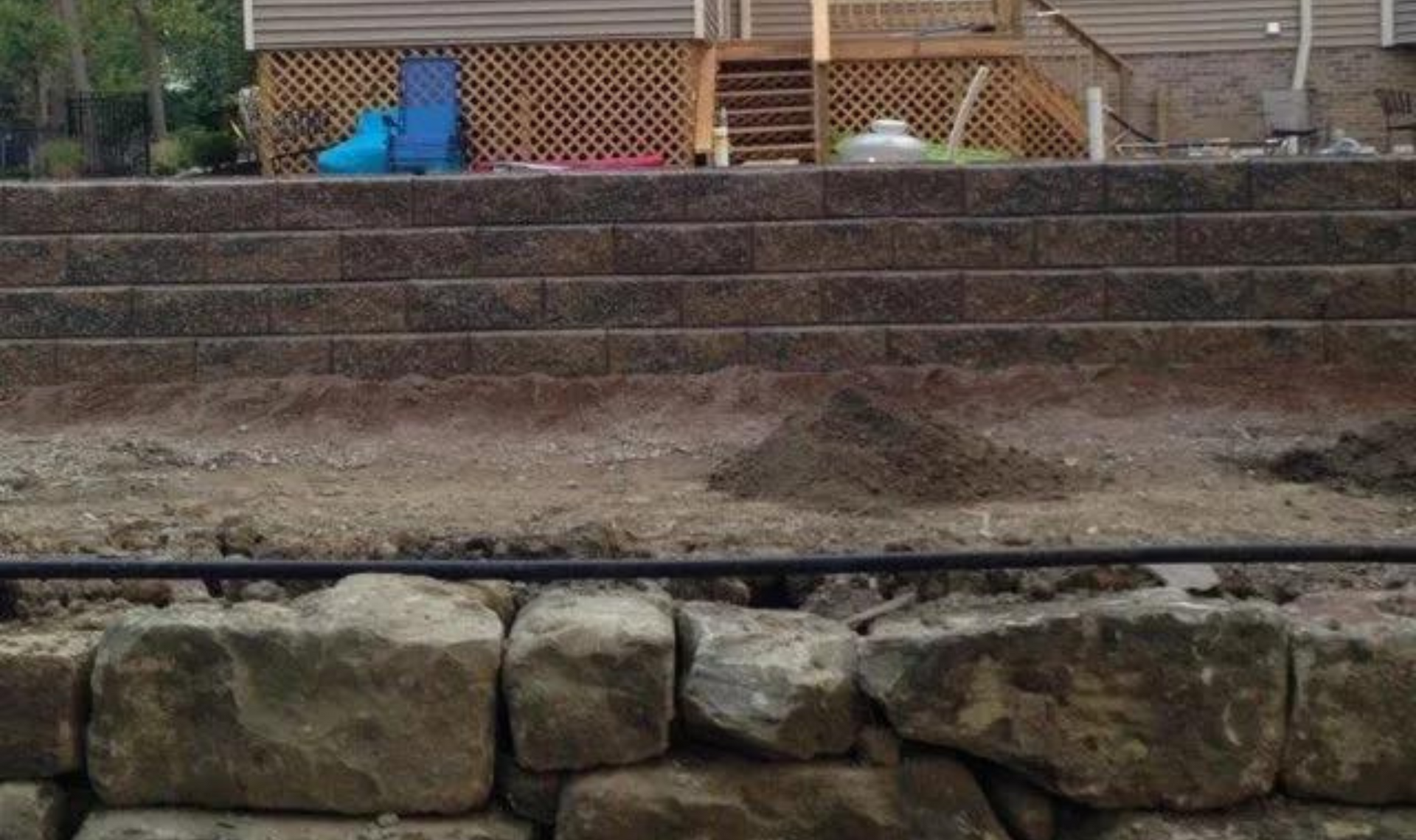 Stone retaining wall and steps leading up to a wooden fence, with a black pipe in the foreground.