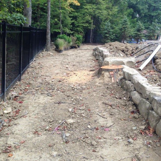Unpaved dirt path beside a black fence and stone retaining wall in a wooded area.
