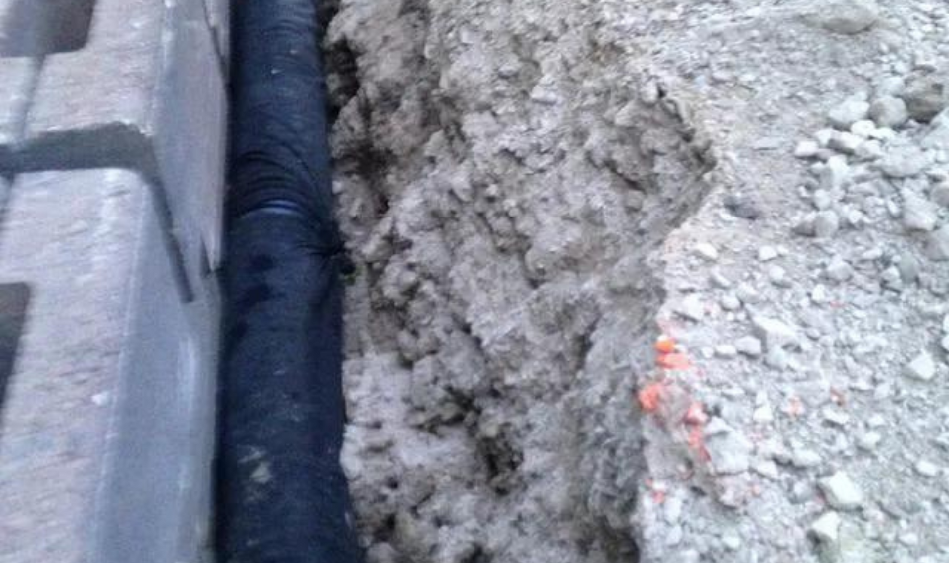 Open trench beside a curb with black pipe and exposed gravel along the edge of a paved road