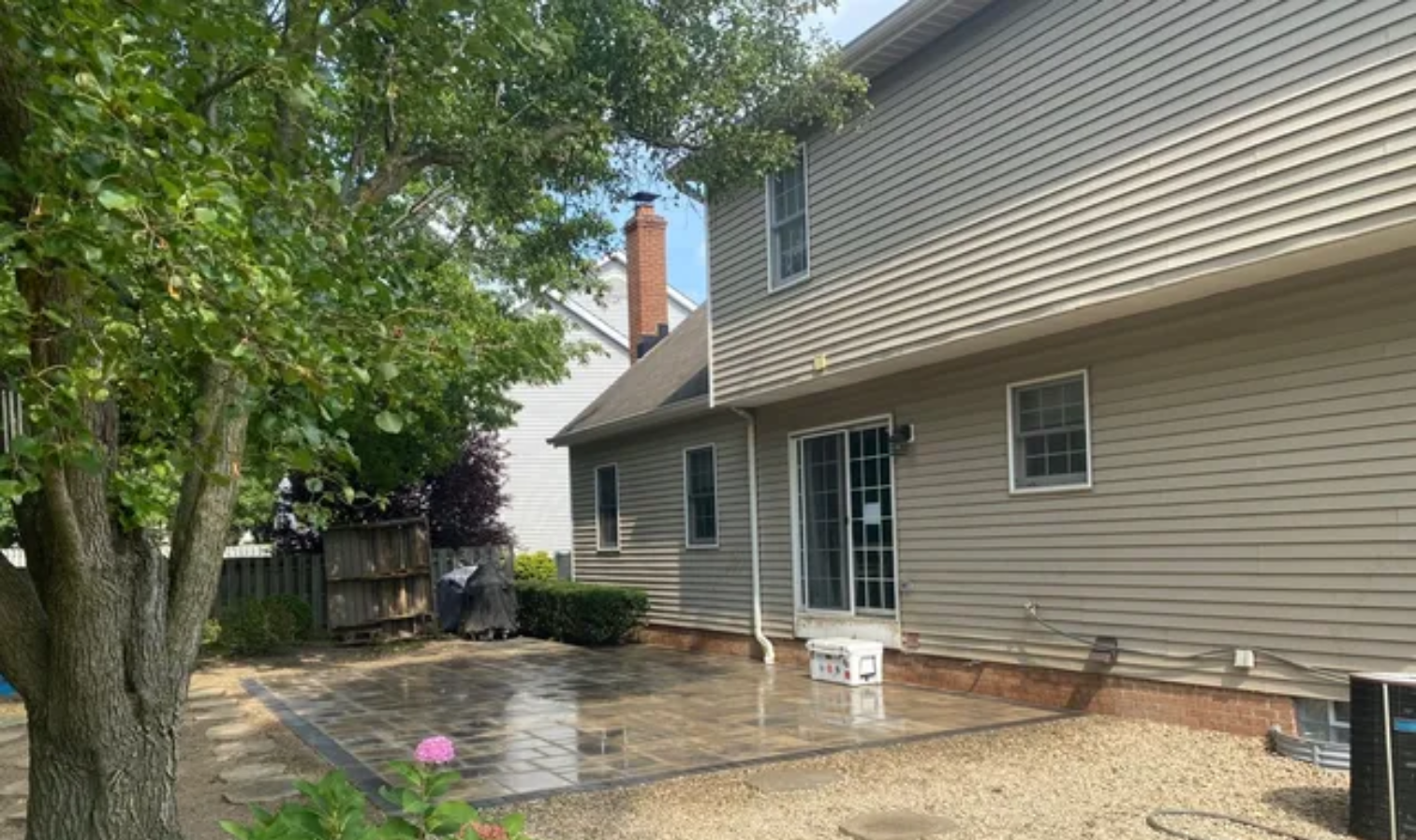 Backyard patio beside beige house with tree, wet concrete, and small flower bed