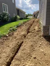 Deep trench dug between two houses along a narrow side yard under a sunny sky