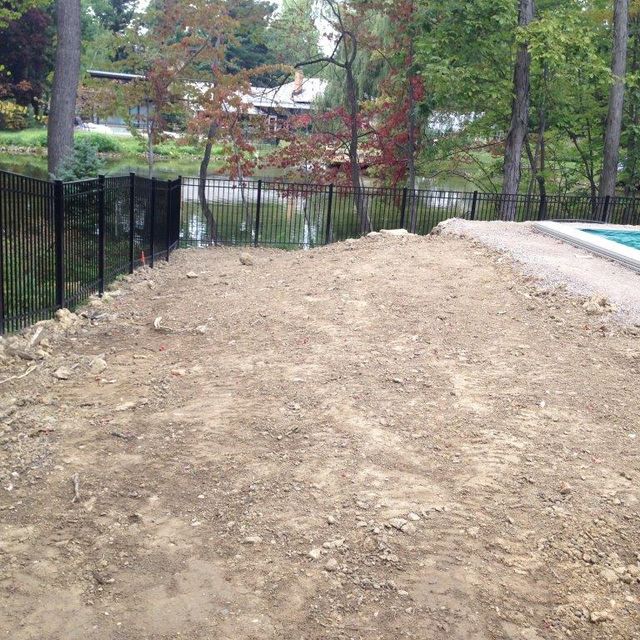 Dirt path under construction beside a black fence and road, with trees and water in the background