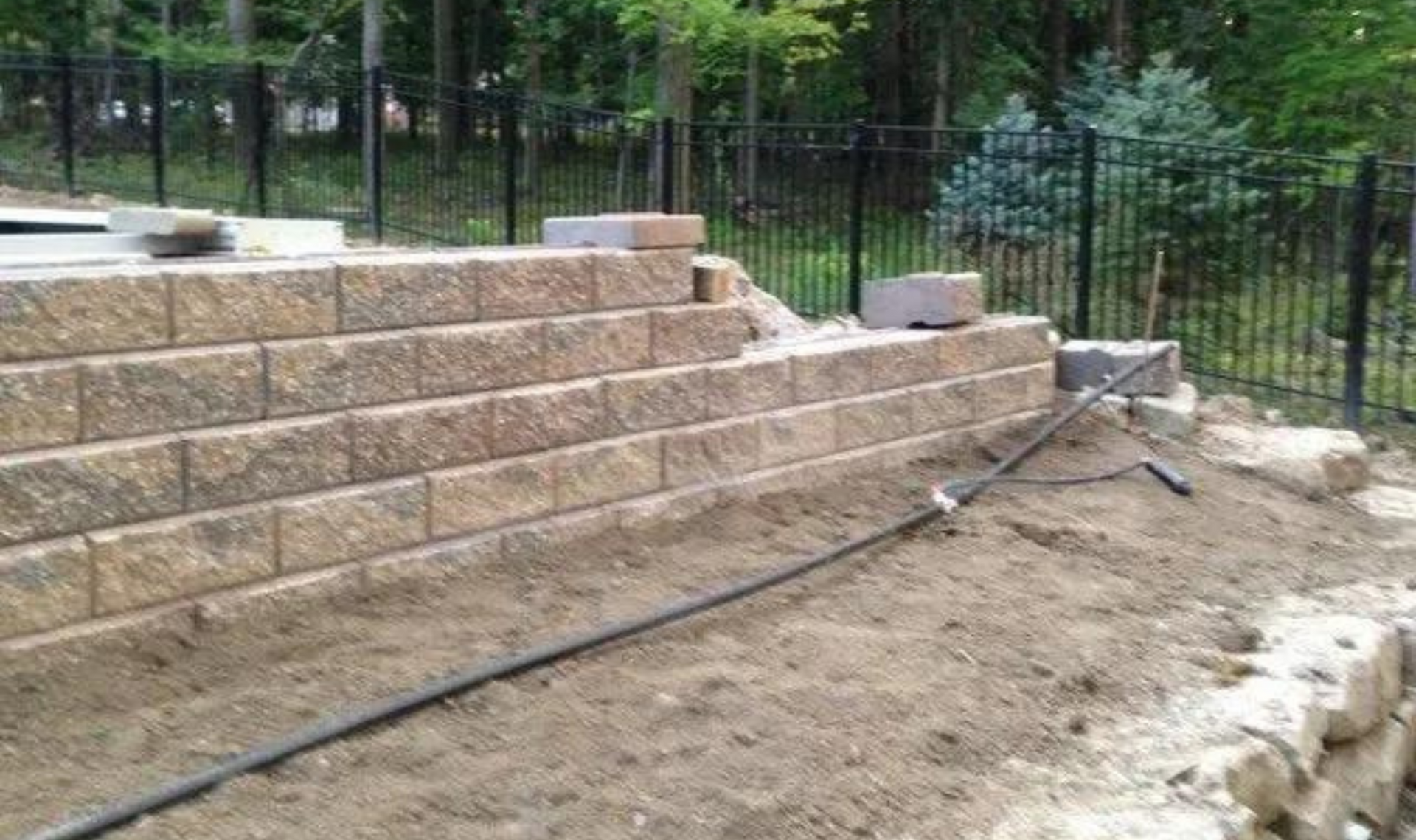 Stone retaining wall under construction beside a dirt slope, with black fencing in the background.