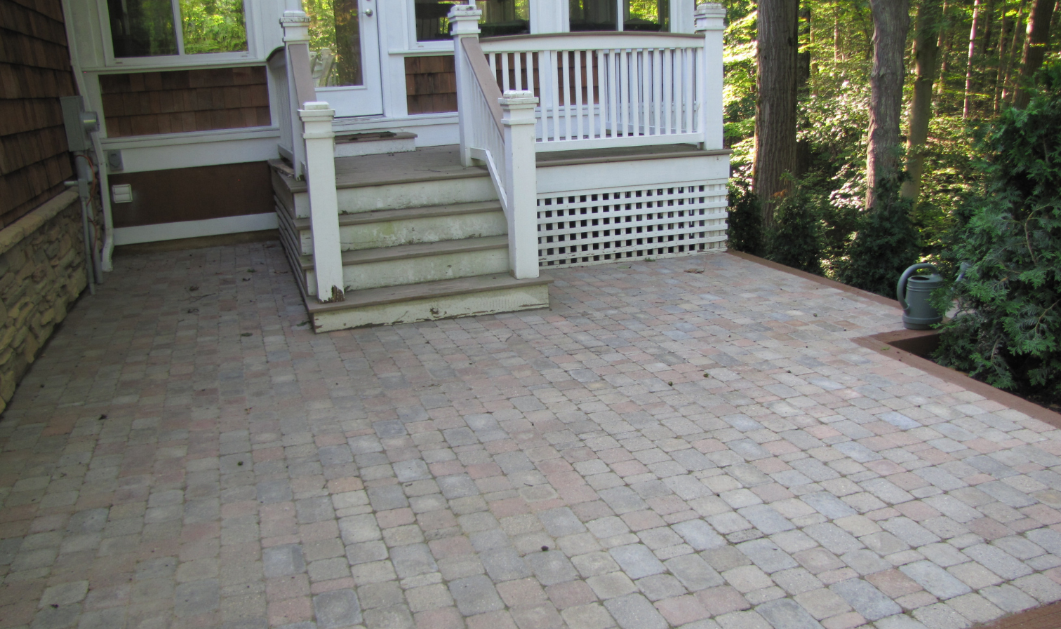 Brick-paved patio leading to white house steps and porch with railing, surrounded by trees