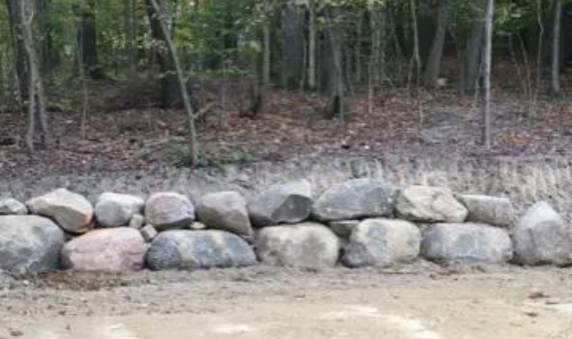 Large stones stacked as a retaining wall in front of a wooded forest edge with a dirt path.