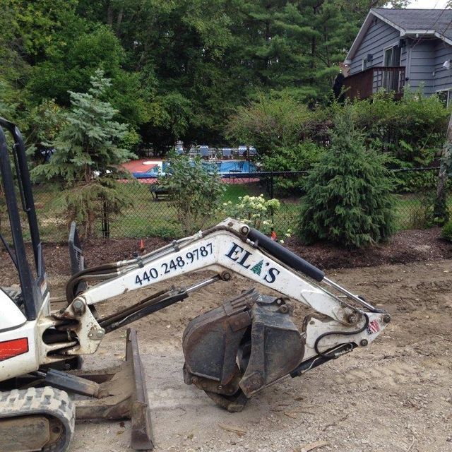 Excavator bucket parked on a dirt path beside a house, with trees and a swimming pool in the background