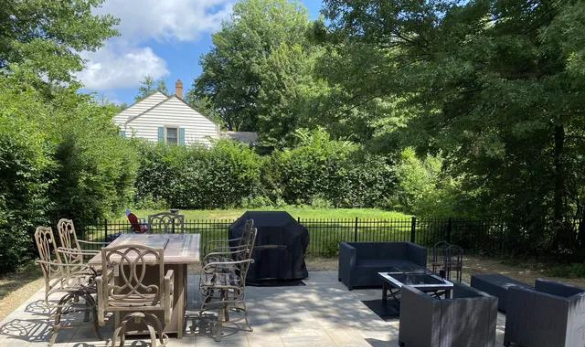Patio with outdoor dining table, grill, and lounge chairs beside a lush green hedge and house