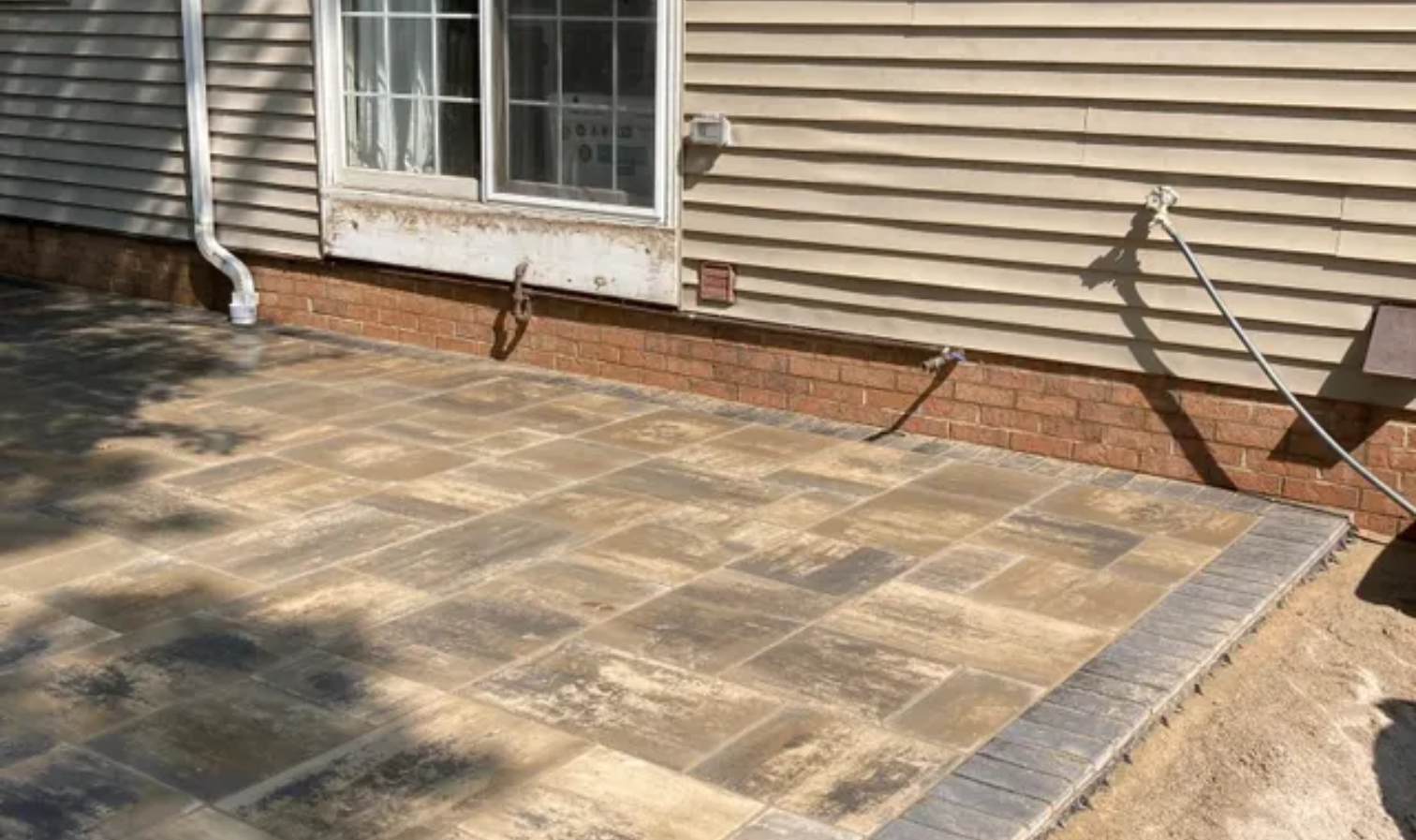 Paver patio beside a house with brick border, window, and downspout in sunlight