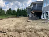 Construction site beside a modern gray house with a dirt lot and trees under a cloudy sky