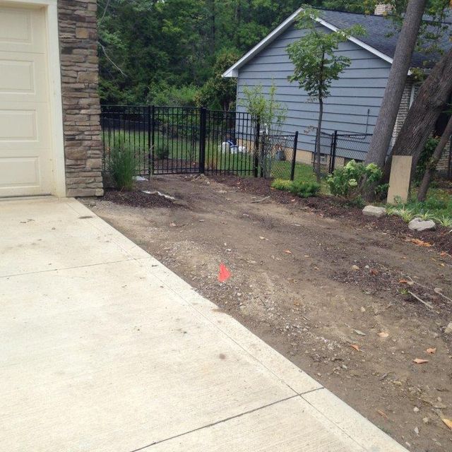 Concrete driveway beside a dirt yard, black fence, and gray shed in a wooded residential area