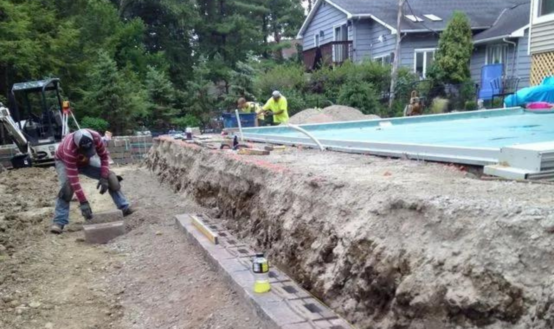 Workers digging a long trench beside a backyard pool at a house under construction