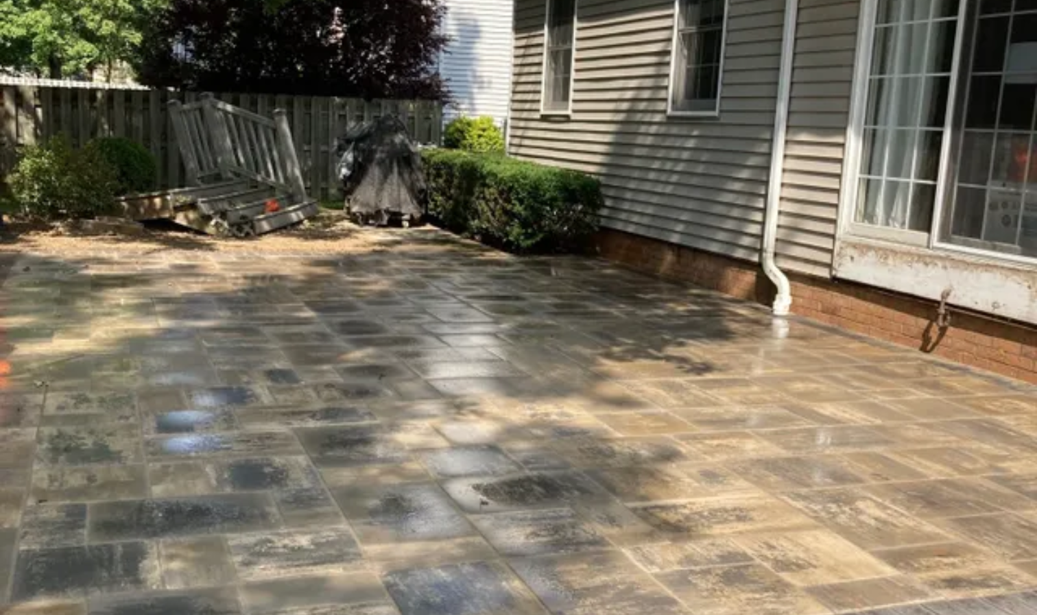 Stone patio beside a house with sliding doors, shrubs, and a fence in the background