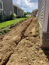 Freshly dug trench running beside a house in a narrow dirt path between residential buildings