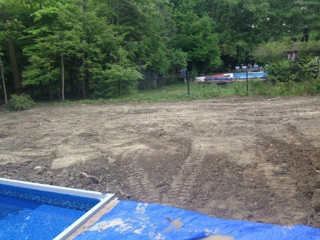 Bare dirt lot beside a blue pool liner, backed by green trees and a parked vehicle in the distance