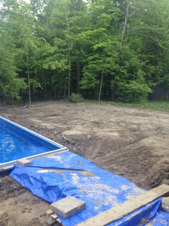 Backyard pool construction site with blue tarp, dirt mound, and trees in the background