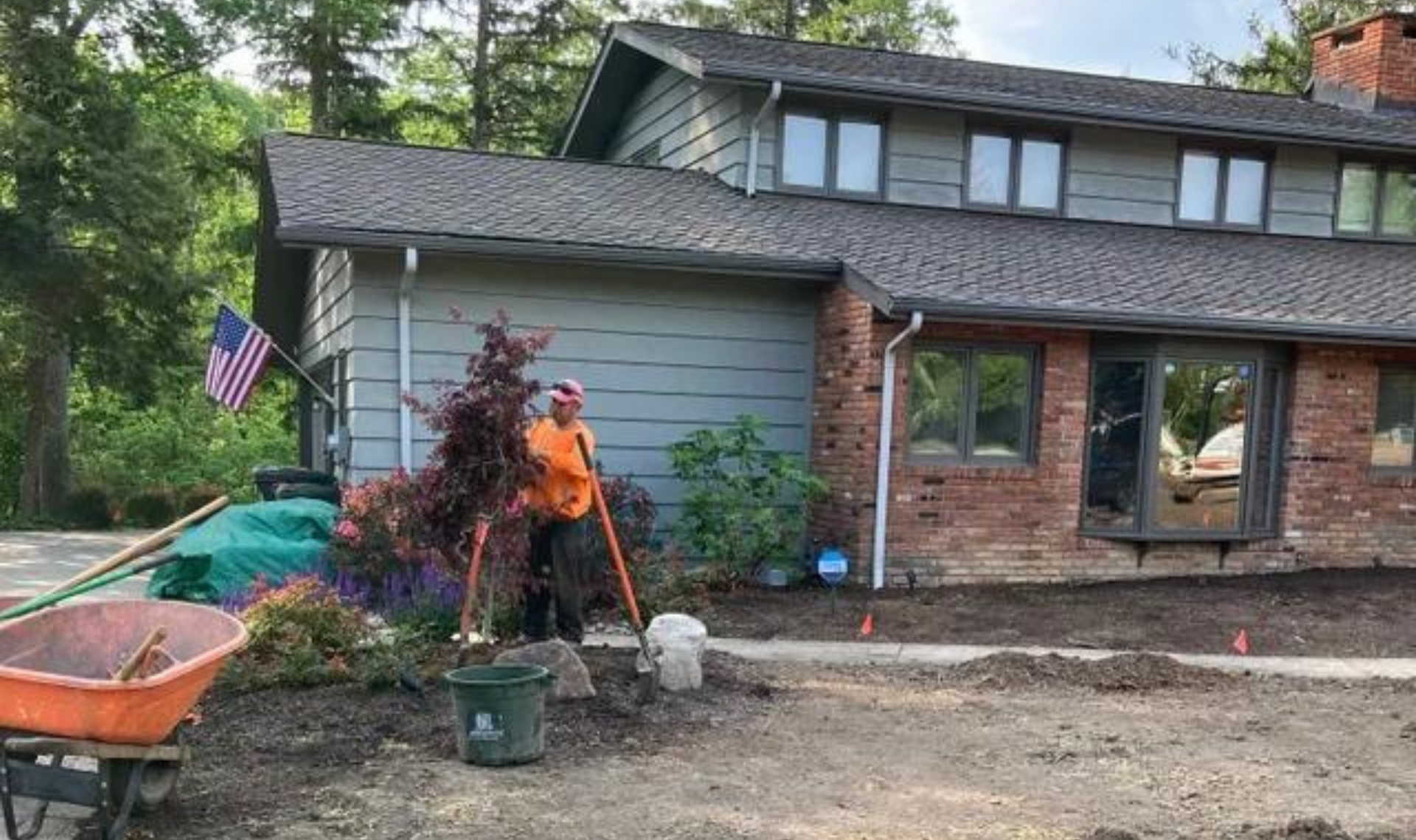 Worker in orange shirt landscaping a front yard beside a brick-and-gray house.