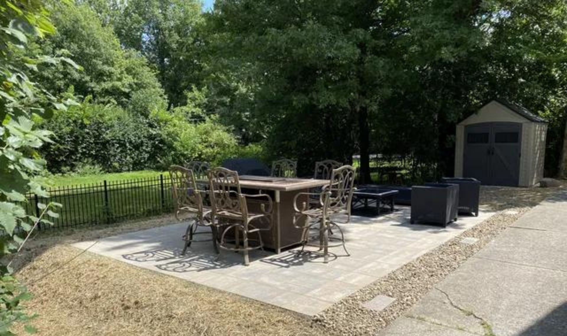 Patio seating area with table, chairs, grill, and shed beside a wooded yard