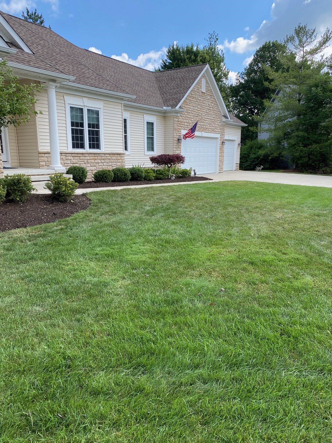 Suburban house with beige siding, white garage, and a green lawn under a partly cloudy sky