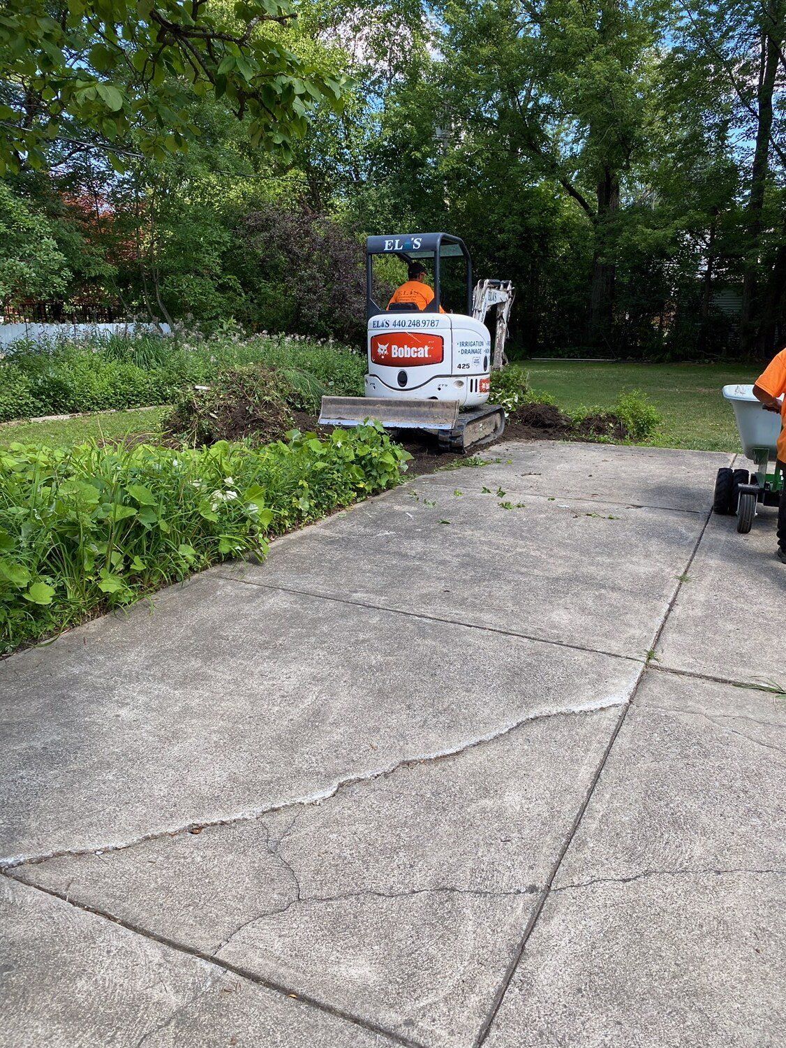 Small construction lift on a driveway near bushes and trees, with a worker beside it.