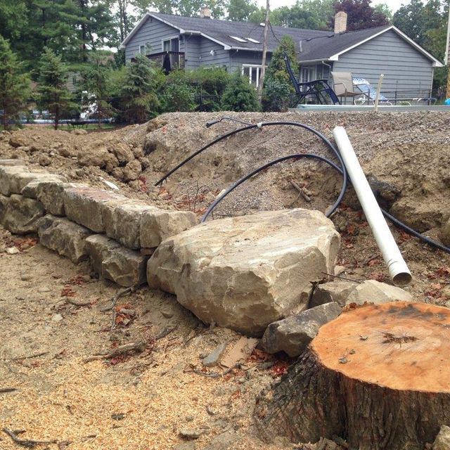 Stone retaining wall under construction in a yard, with dirt, pipes, and a cut tree stump in front of houses.