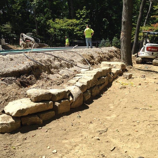 Stone retaining wall under construction in a wooded area, with workers and a truck nearby