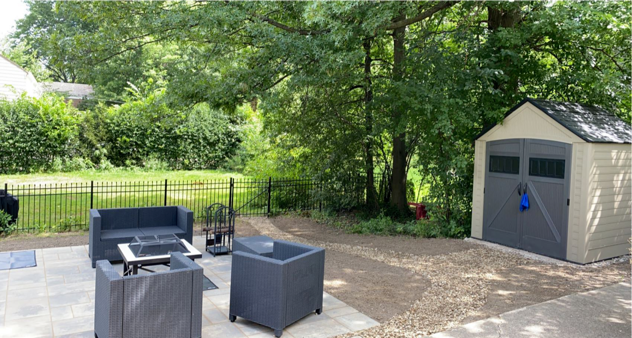 Backyard patio with gray wicker chairs, fire pit, and a beige storage shed under trees