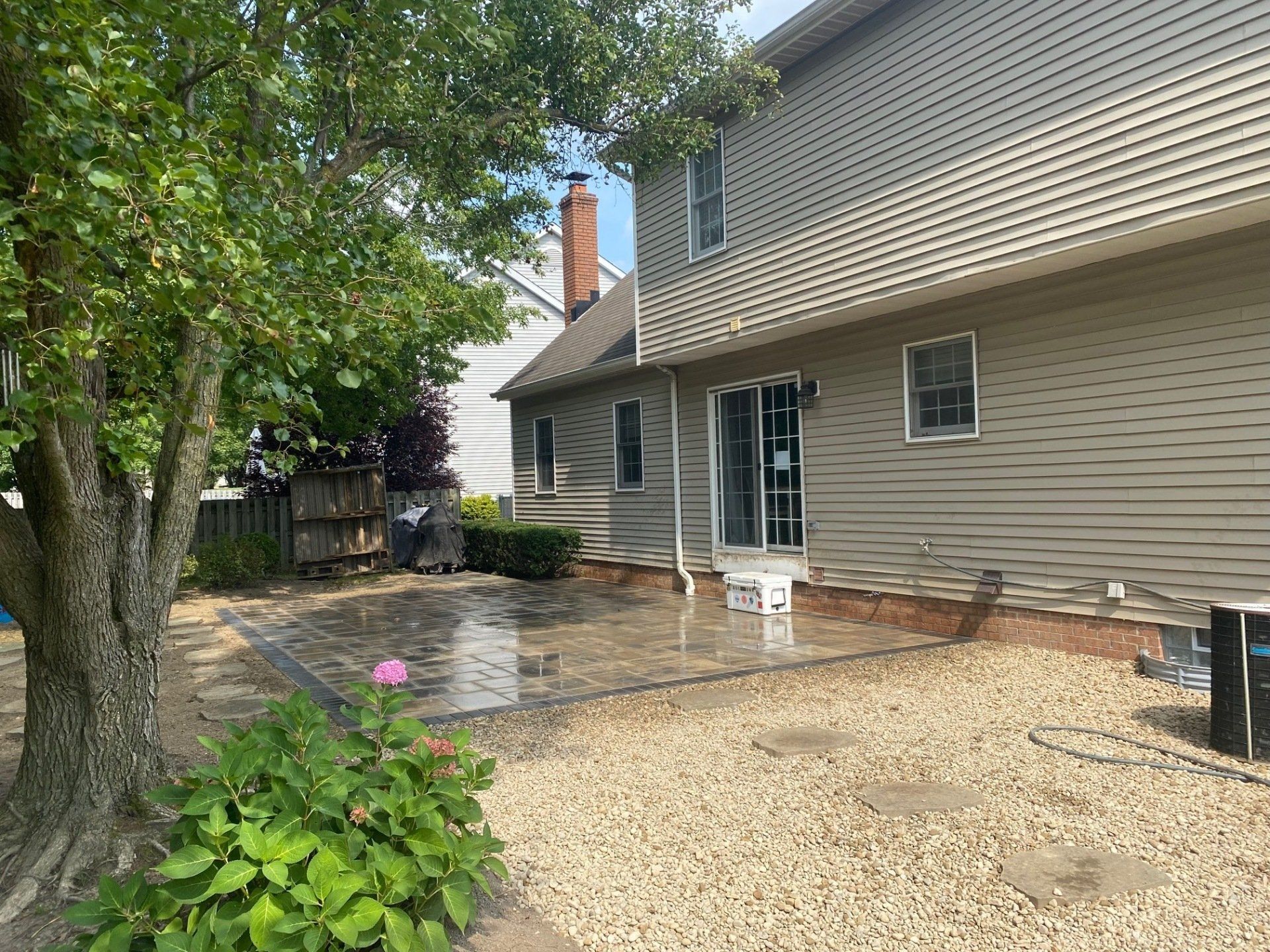 Backyard patio with wet concrete, gravel path, tree, and beige house siding