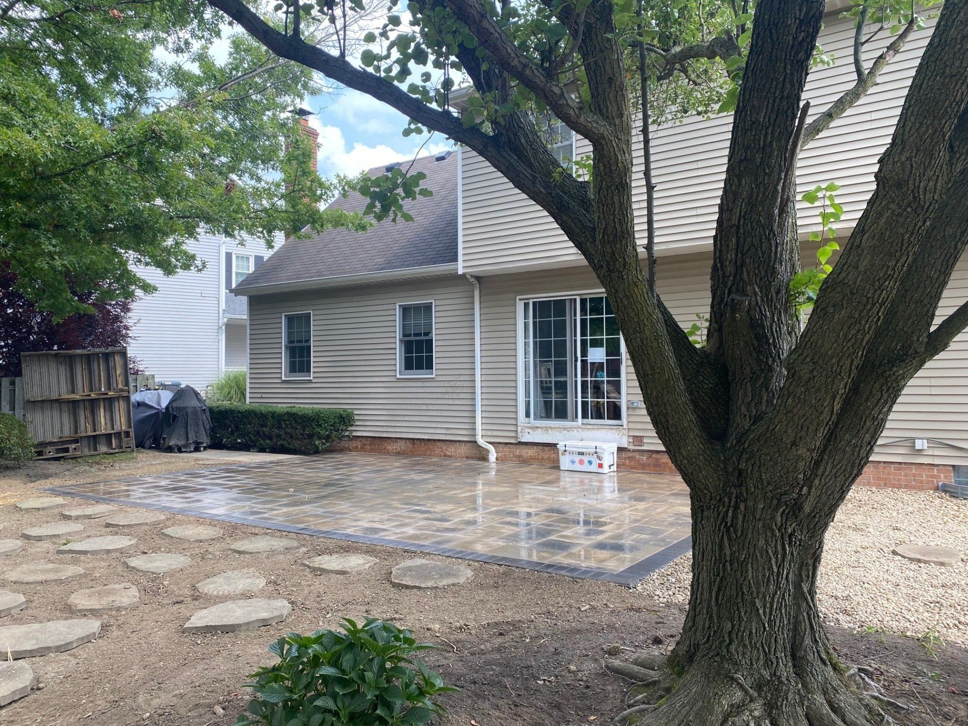 Backyard patio with stone pavers, a large tree, and a beige house with sliding glass doors
