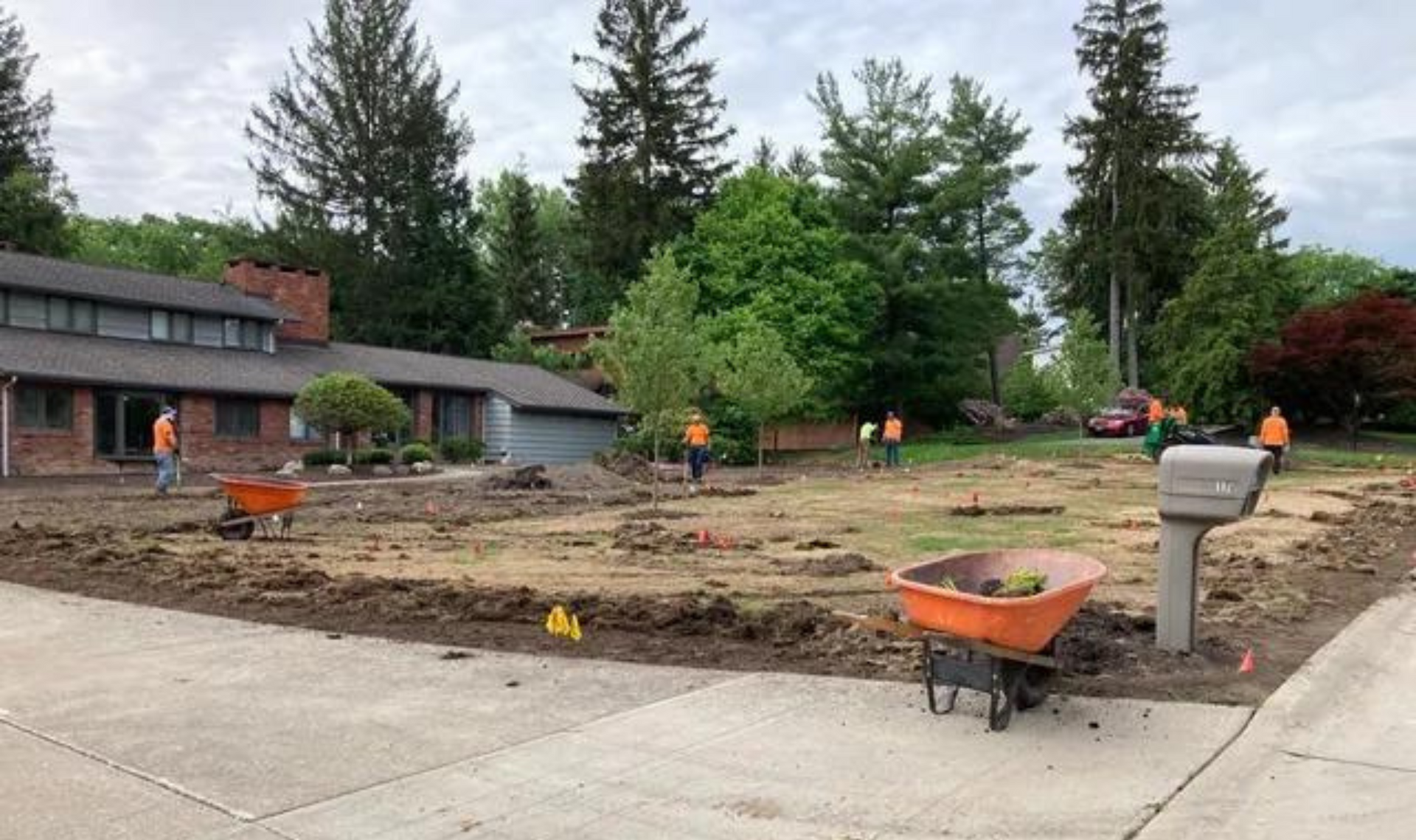 Construction workers leveling dirt in a front yard beside a house, with wheelbarrows and tools.