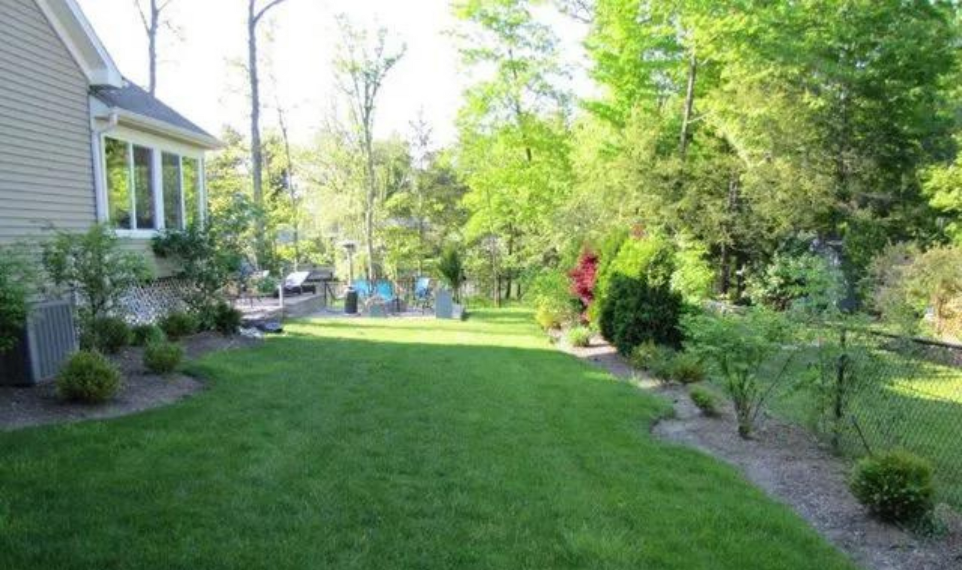Green backyard lawn beside a house, with trees, shrubs, and a patio area in bright sunlight.