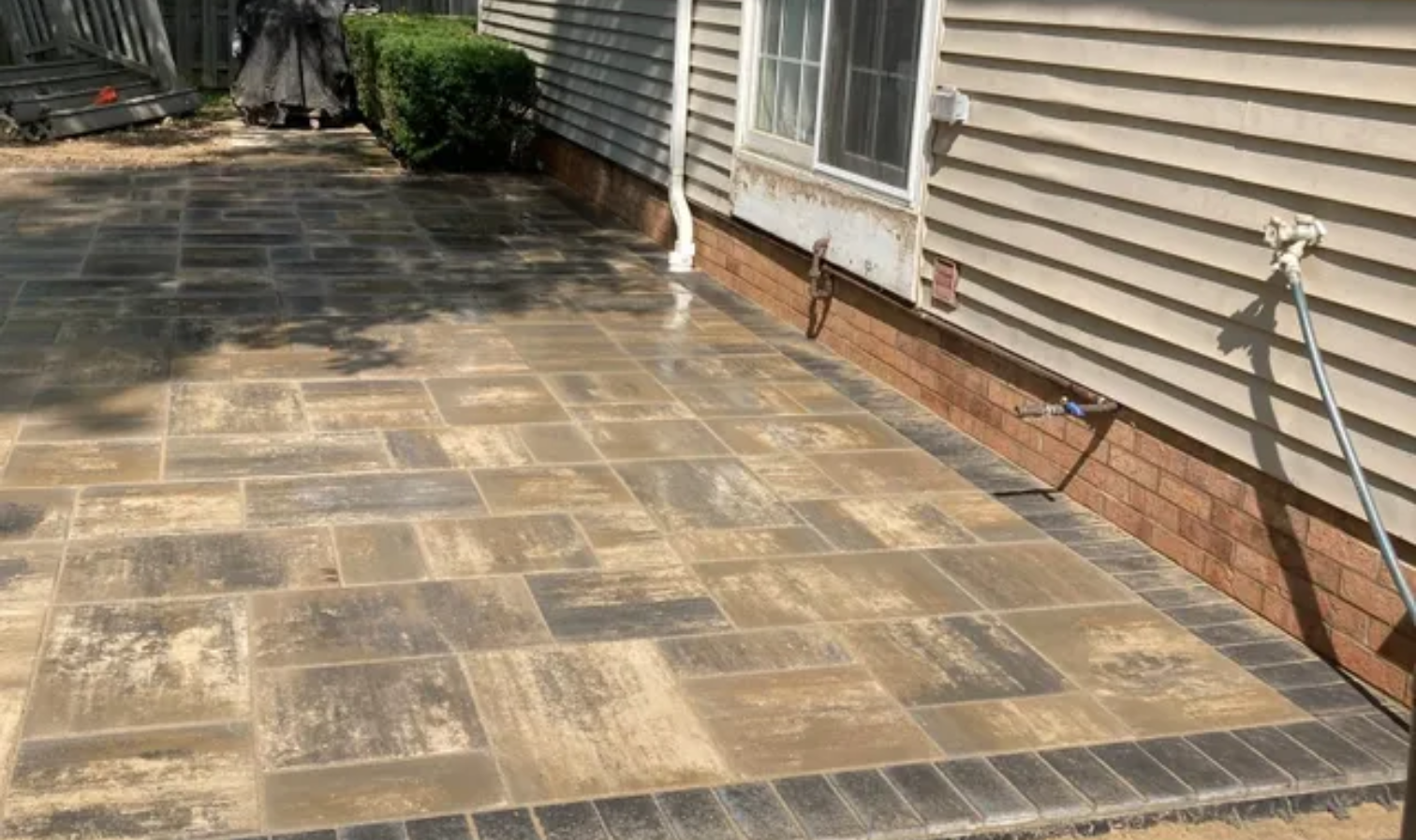 Stone patio beside a house with sliding glass doors and tan siding, bordered by a brick foundation.