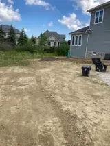 Partially graded dirt lot beside a gray house with a few black bins and other homes in the background