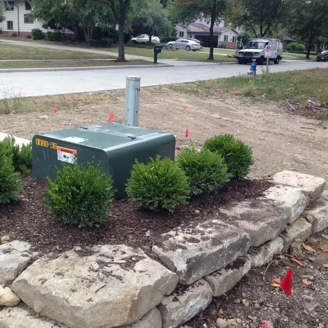 Utility box beside landscaped stone retaining wall in a suburban yard, with red survey markers nearby