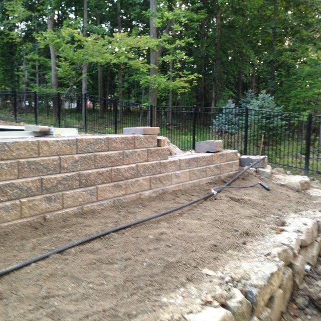 Outdoor stone retaining wall under construction beside a wooded yard, with exposed soil and black fencing.