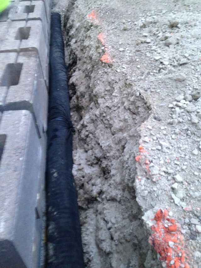 Narrow trench between gray pavers and rough dirt ground with a black drainage pipe running along the bottom