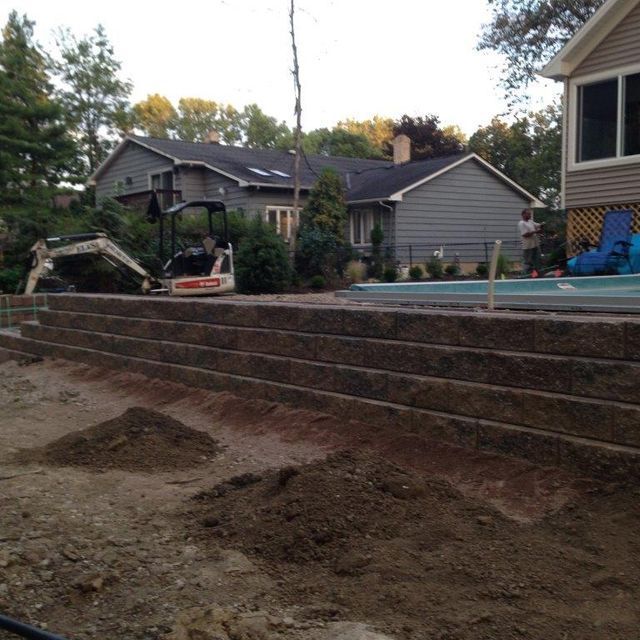 Excavator grading a dirt yard beside a house with terraced retaining walls and a pool in the background