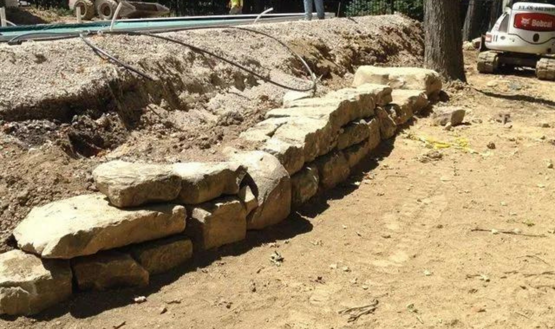 Stone retaining wall under construction along a dirt path, with rocks and a tree nearby