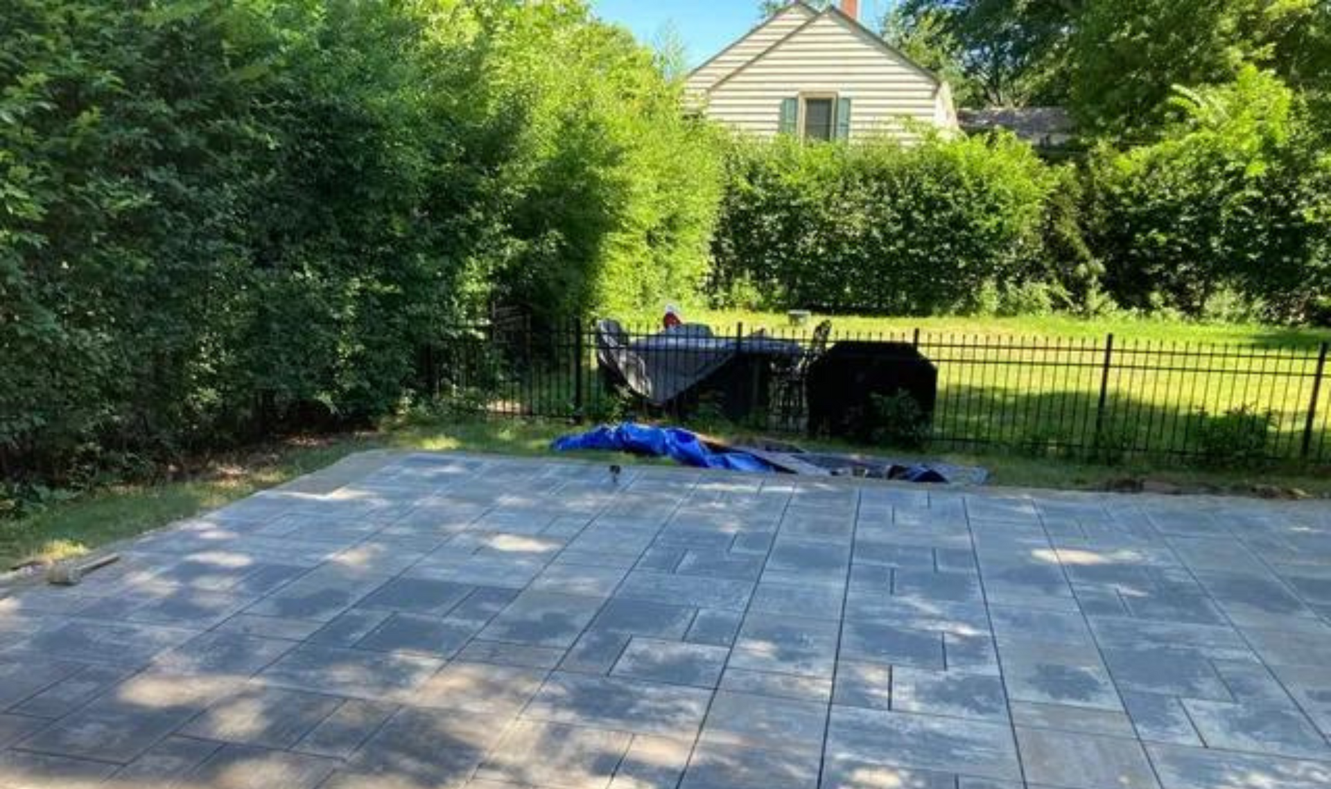 Patio with gray pavers, black fence, blue tarp, and dense green hedges in a backyard setting