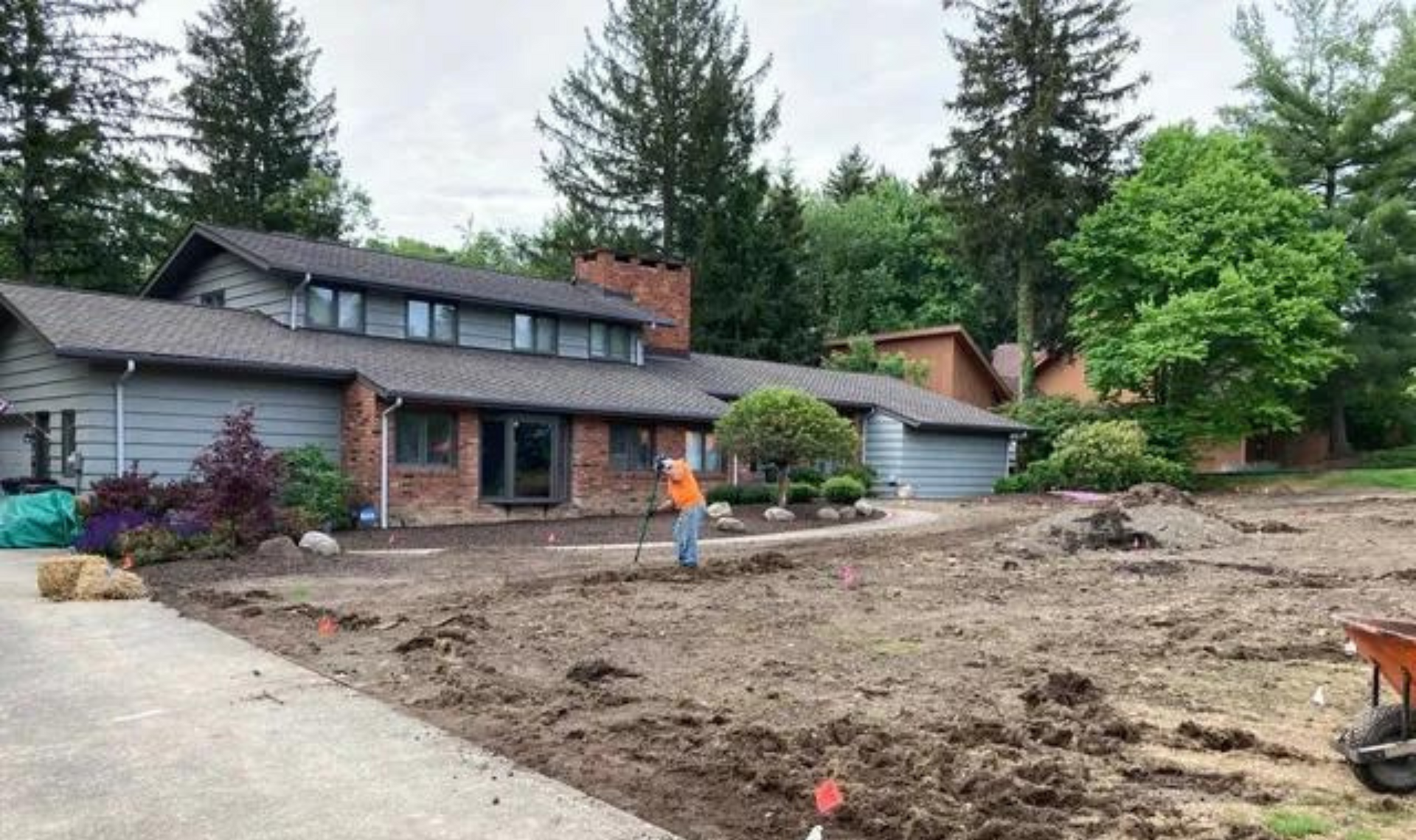 House under renovation with torn-up front yard, worker in orange vest, and trees in the background