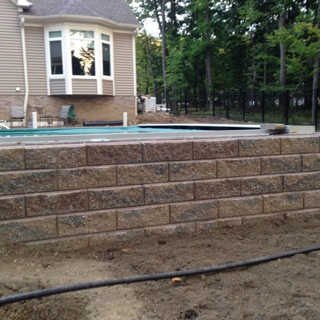 Backyard pool with stone retaining wall behind a tan house, surrounded by trees and a black fence.