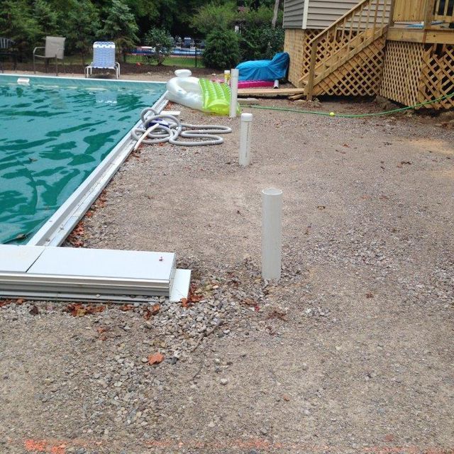 Swimming pool edge beside a gravel patio with coiled hose, lounge chair, and wooden deck steps