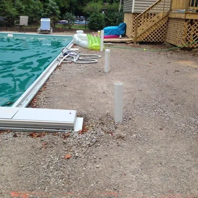 Poolside gravel area with white posts, coiled hose, and a wooden deck in the background
